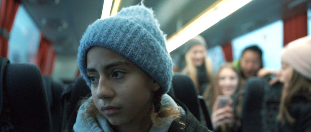 En jente sitter lei seg på bussen helt alene. Bak henne sitter det flere jenter og ler av henne og tar bilder av henne med mobiltelefonen.