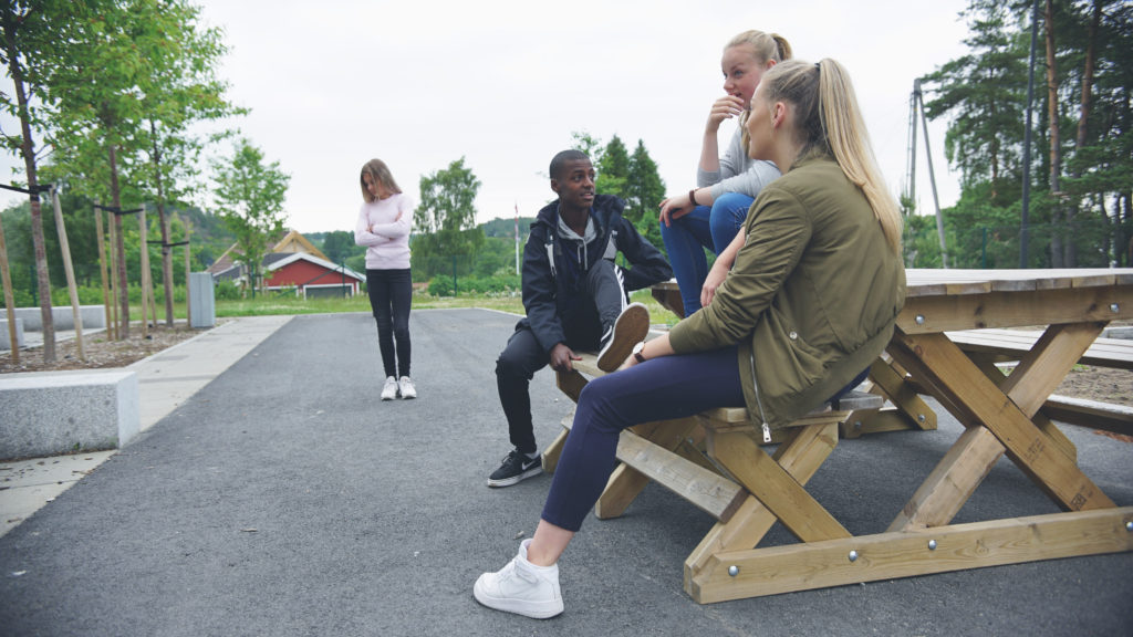 Tre ungdom sitter på en benk, mens det står en annen jente alene. Hun føler seg utenfor.