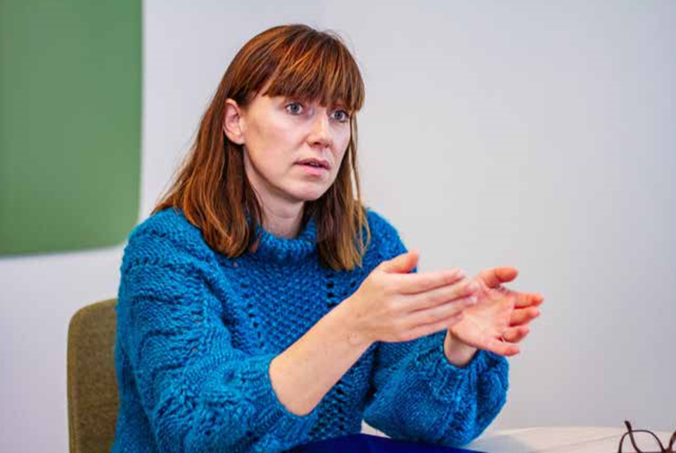 Åse har blå genser og sitter i et rom og gestikulerer