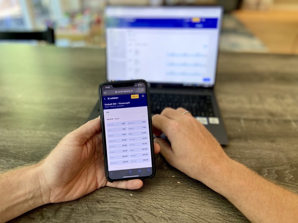En mann holder en telefon med oddsen fra Norsk Tipping. Han har tilsvarende skjerm på laptop