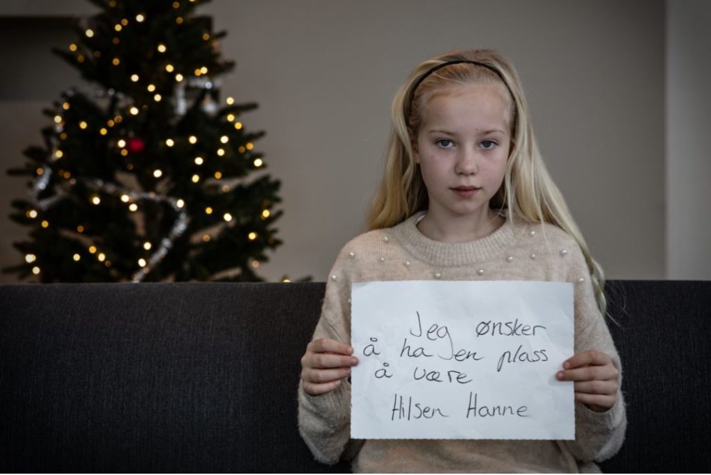 Jente med langt lyst hår sitter i et rom med juletre og med en lapp der det står "Jeg ønsker meg en plass å være"