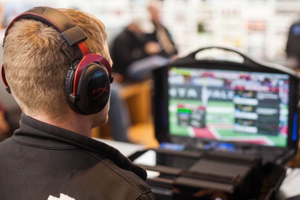 Mann ser på gaming på sin PC med et stort headset på hodet. Det er en annen person som sitter i bakgrunnen