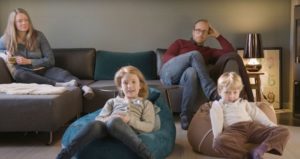 To foreldre i hvert sitt hjørne av en sofa og to barn i saccosekker foran på gulvet