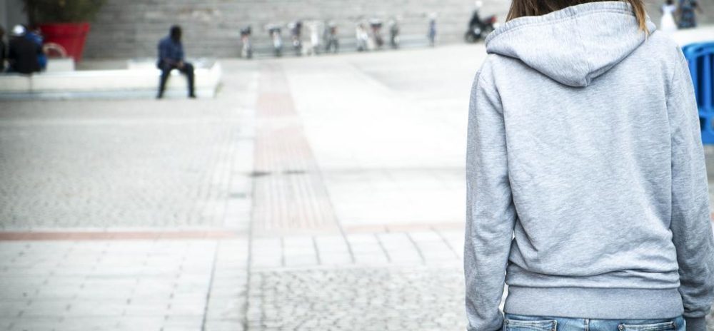 Jente med ryggen til kledd i grå hettegenser ser utover en tom, grå plass. Ett menneske og noen sykler i bakgrunnen