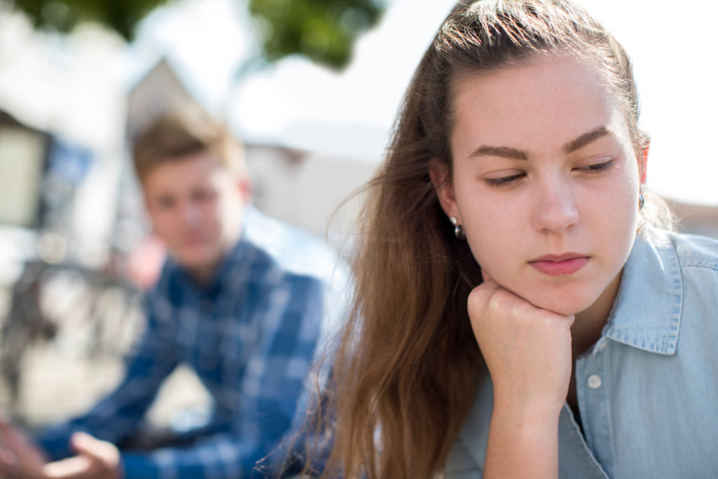 En tenåringsjente sitter med et bekymringsfylt uttrykk i ansiktet. Bakgrunnen er uklar, men man skimter så vidt broren som sitter bak henne.