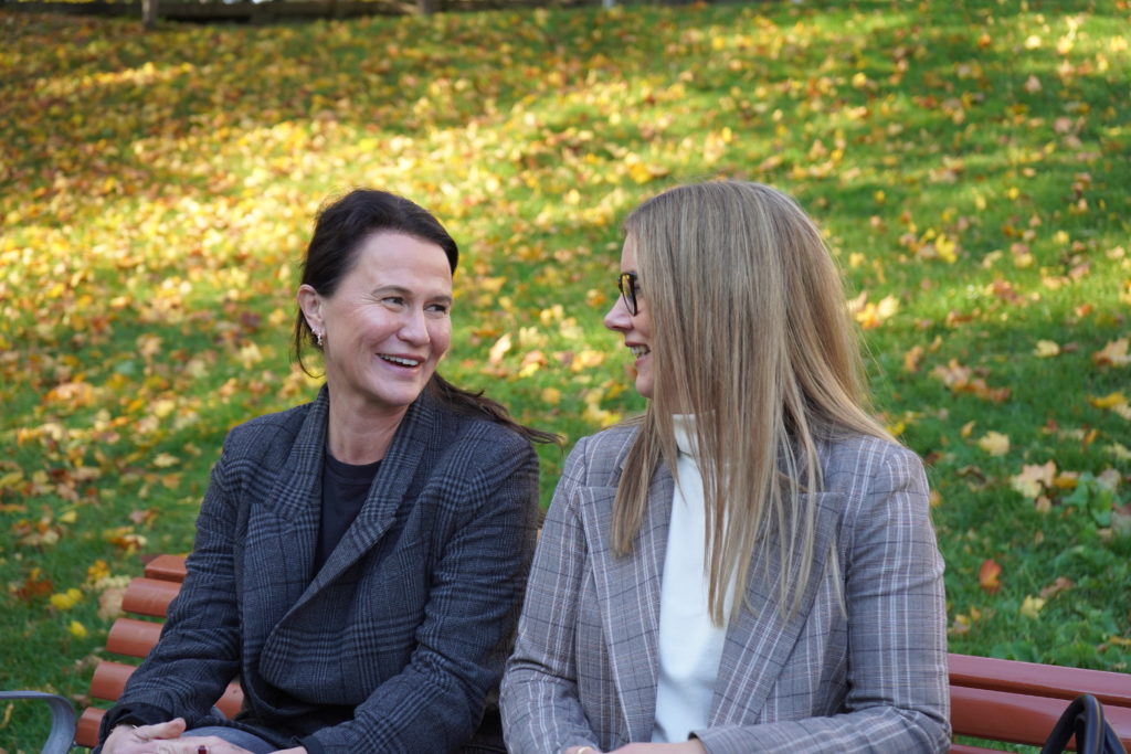 Camilla Dahle og Trine Ulvang ser på hverandre og smiler mens de sitter på en benk i strålende høstvær
