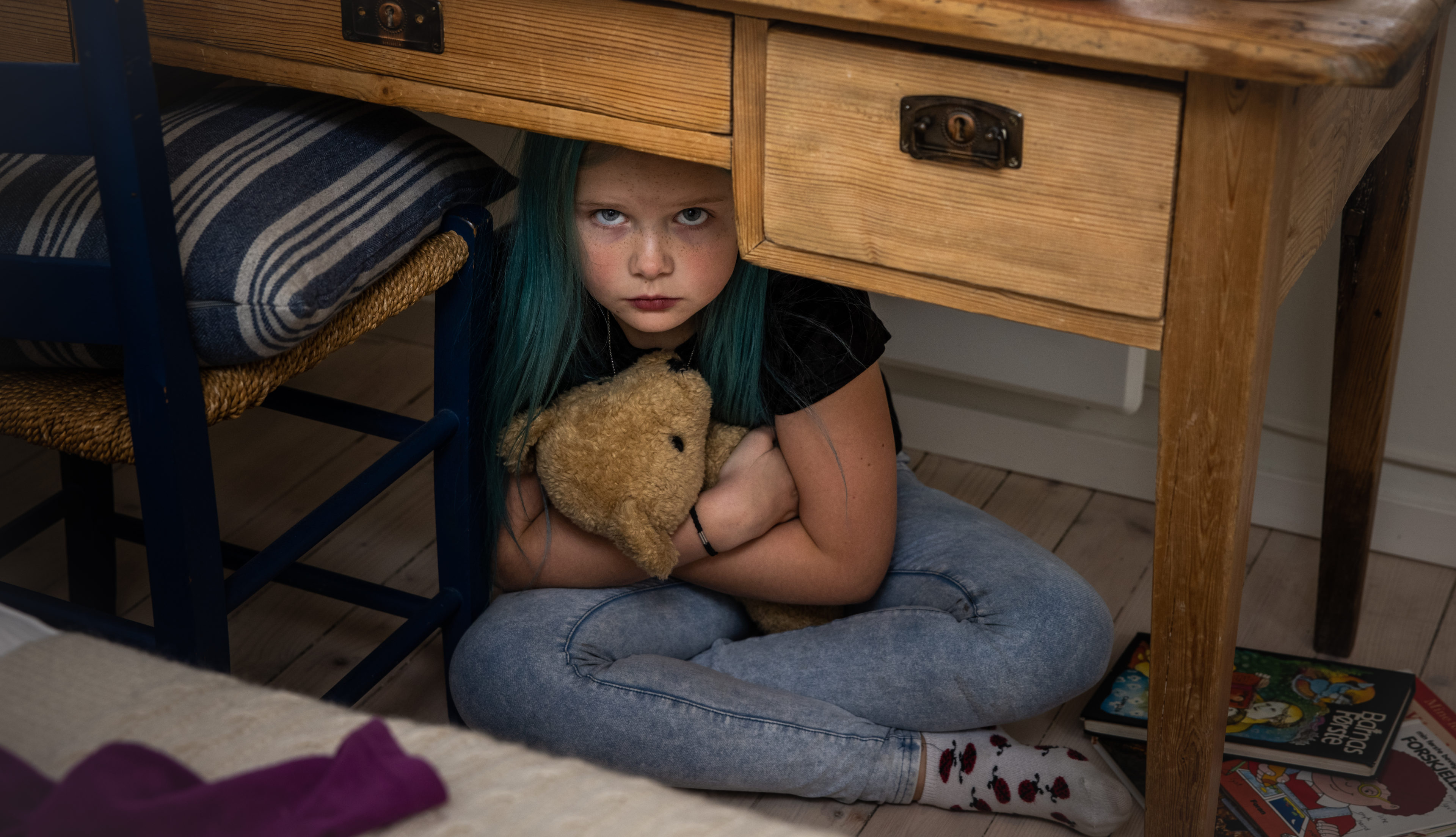En ung jente med blått hår gjemmer seg under pulten med en beskyttende arm rundt bamsen sin mens hun stirrer alvorlig opp mot kameraet.