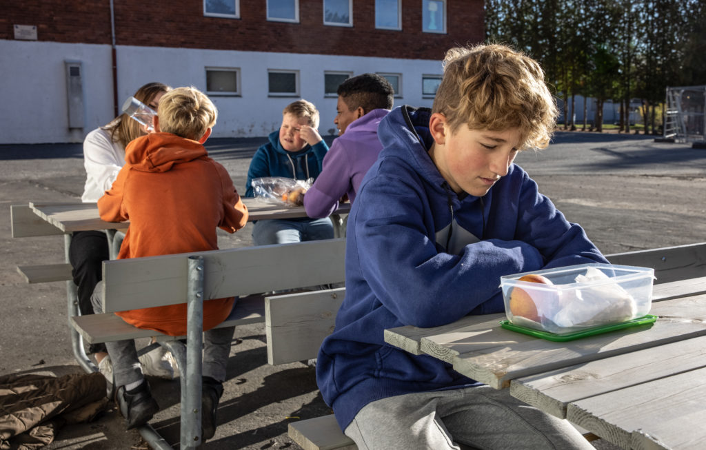 En gutt føler seg utenfor i skolegården. Han sitter alene med lunsjen mens det sitter 4 andre bak han