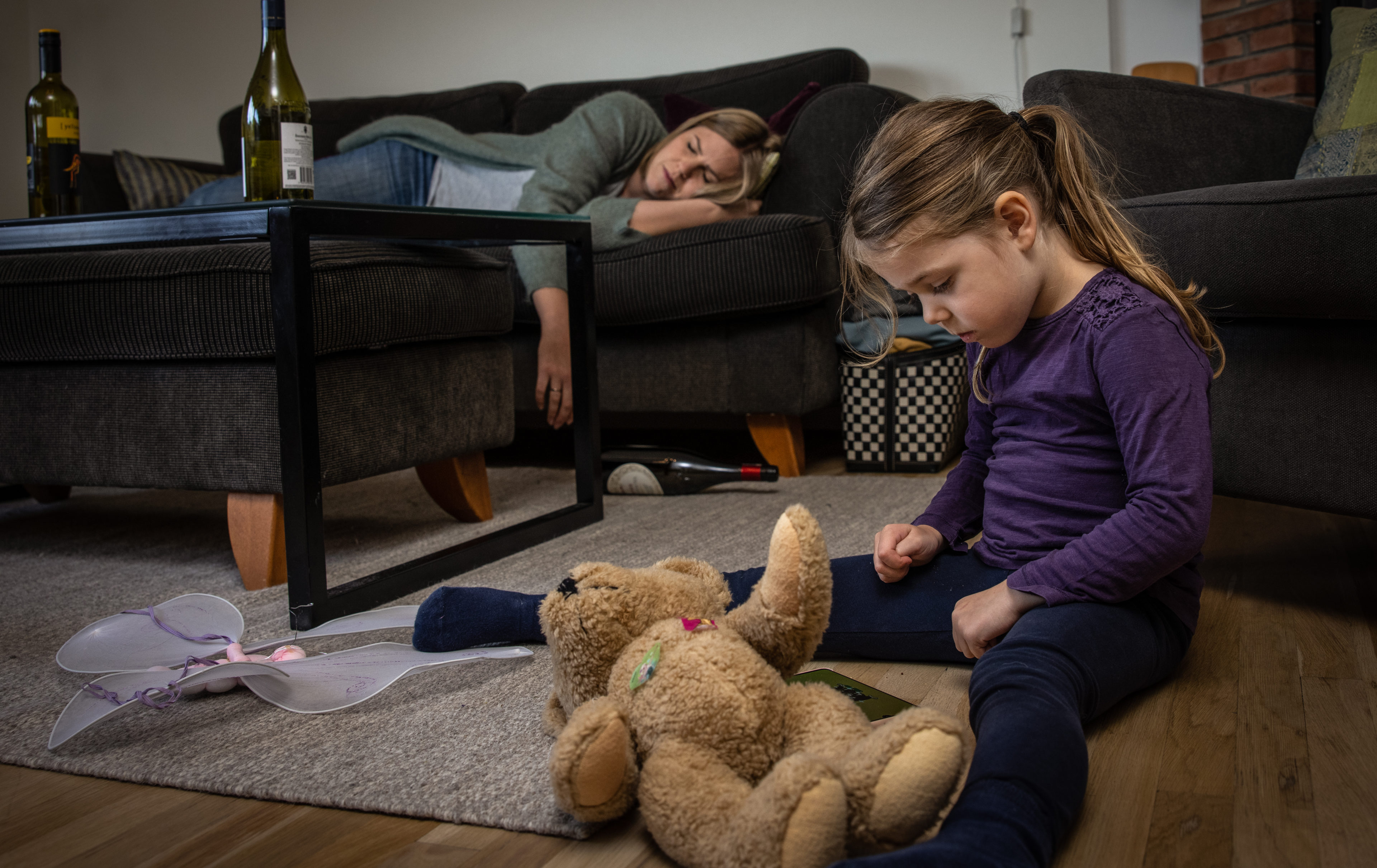 En jente sitter En jente sitter på gulvet og ser ned på bamsen sin. Hun er lei seg. Moren hennes ligger og sover på sofaen og man ser vinflasker på bordet
