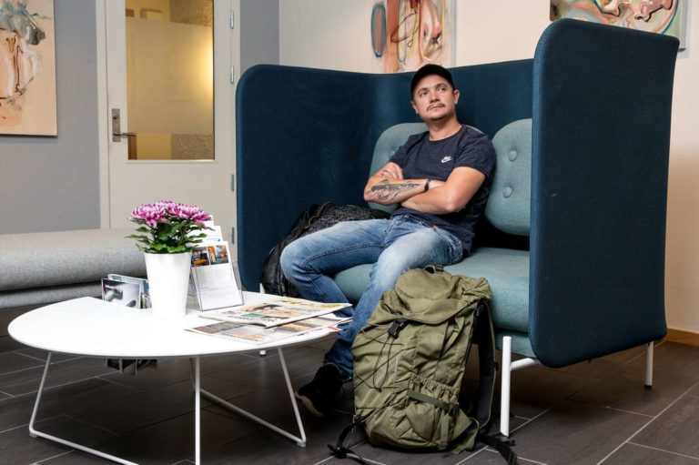 En mann sitter på venterommet med armer i kors. Det er et innbydende rom med en grønn sofa, blomst på bordet og kunst på veggene