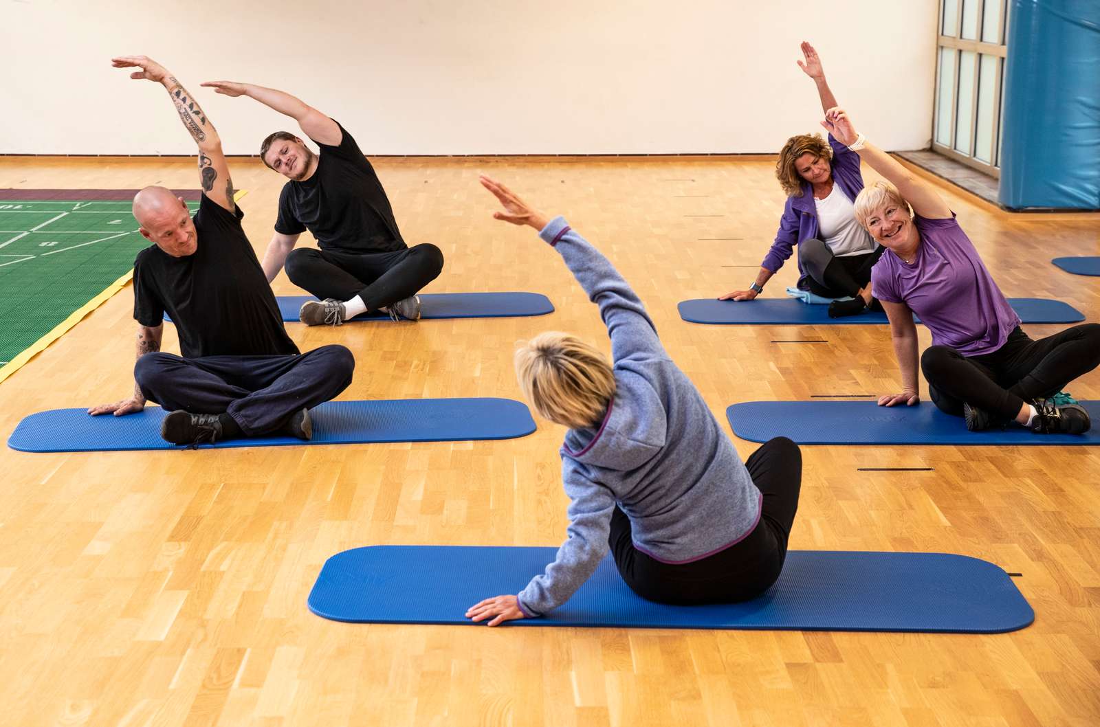 Det er fem mennesker som trener yoga på matter. To menn og tre damer, og de strekker armene over den ene siden.