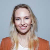 Kvinne med lyst hår, hun smiler, og har på seg en oransje jakke med en hvit bluse under