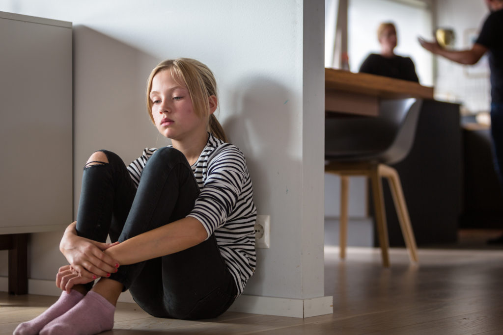 En ung jente som sitter på gulvet og er lei seg for at foreldrene krangler i bakgrunnen