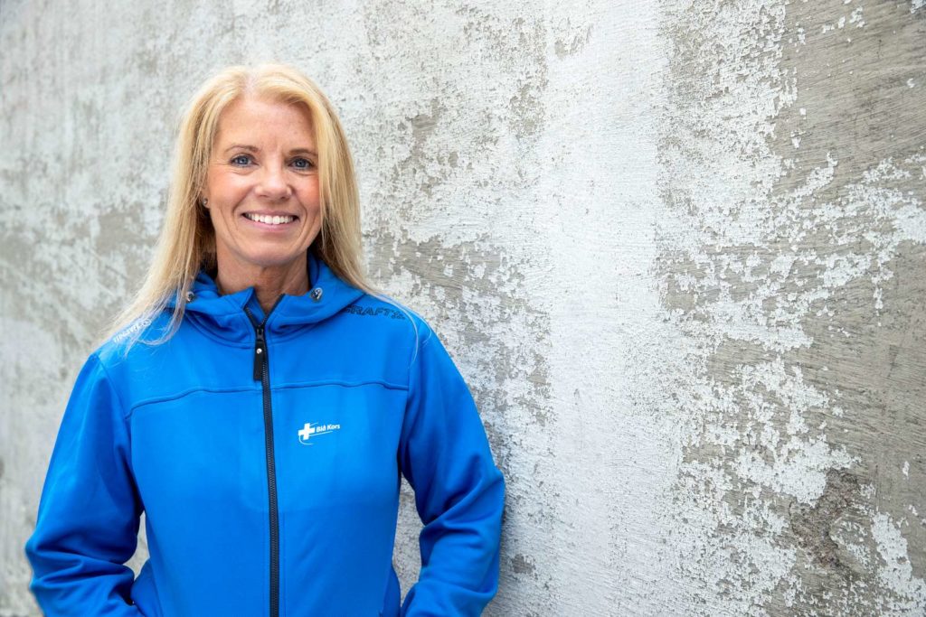 Trine Stensen står i blå jakke med Blå Kors logo. Hun smiler foran en murvegg