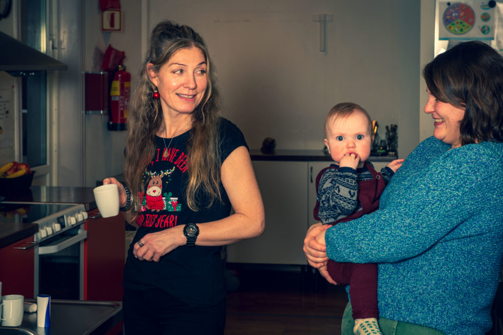To kvinner og et lite barn på 1 år er inne på et kjøkken. Kvinnene ser på hverandre og smiler. Dene ene kvinner har en kaffekopp i hånden