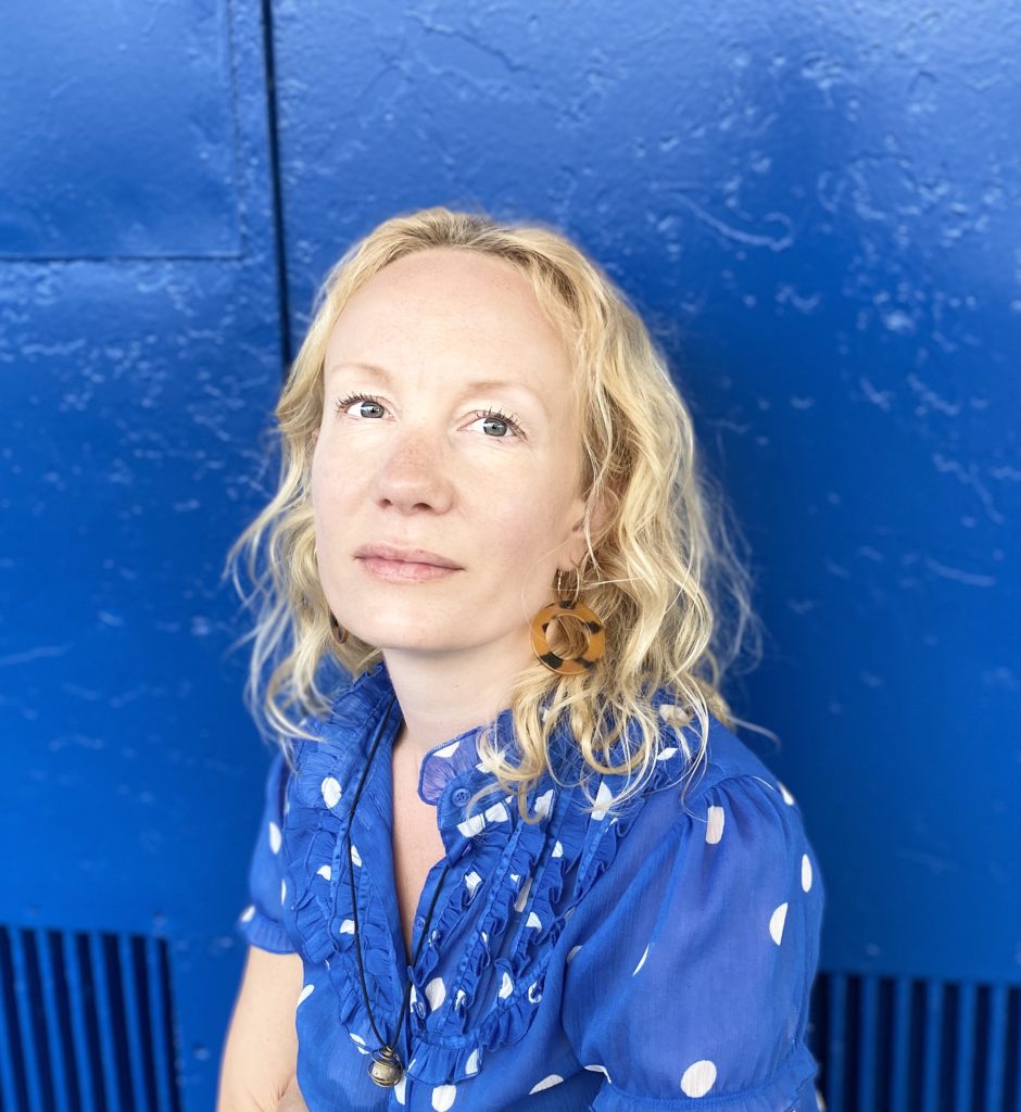 Hanne Backe-Hansen sitter i blå bluse som matcher perfekt den blå veggen hun sitter foran