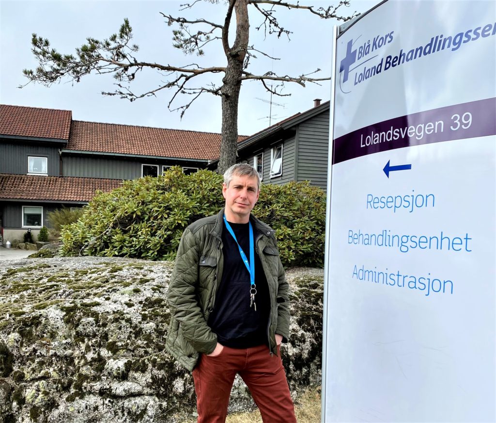 Frode Holmen står foran velkomstskiltet til Blå Kors klinikk, Loland med hendene i lomma og et smil om munnen.