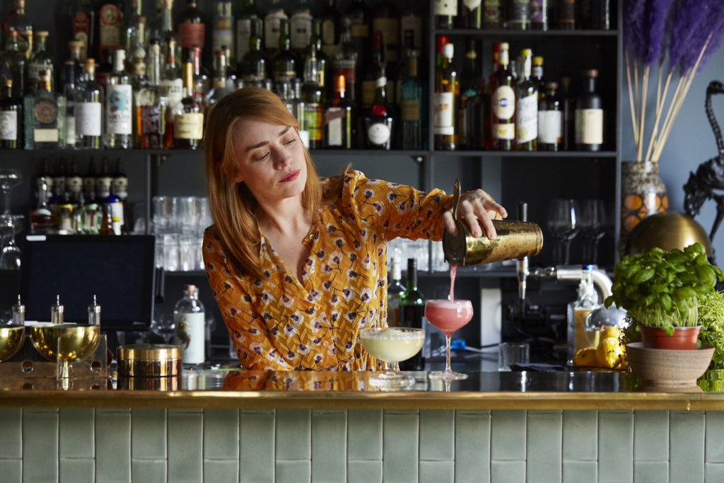 En kvinnelig barntender heller i drikke i to glass som står på bardisken. Det er to alkoholfrie drinker