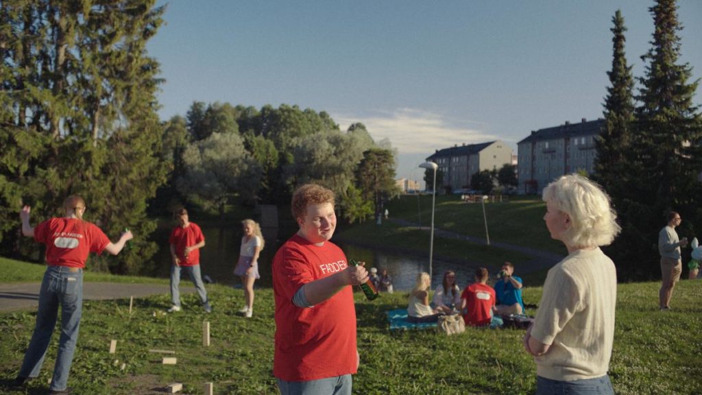 En ung mann som er student spør en annen student om hun vil ha øl. Han rekker ut ølen. Han har rødt hår, og har en rød t-skjorte på seg som det står fadder på.