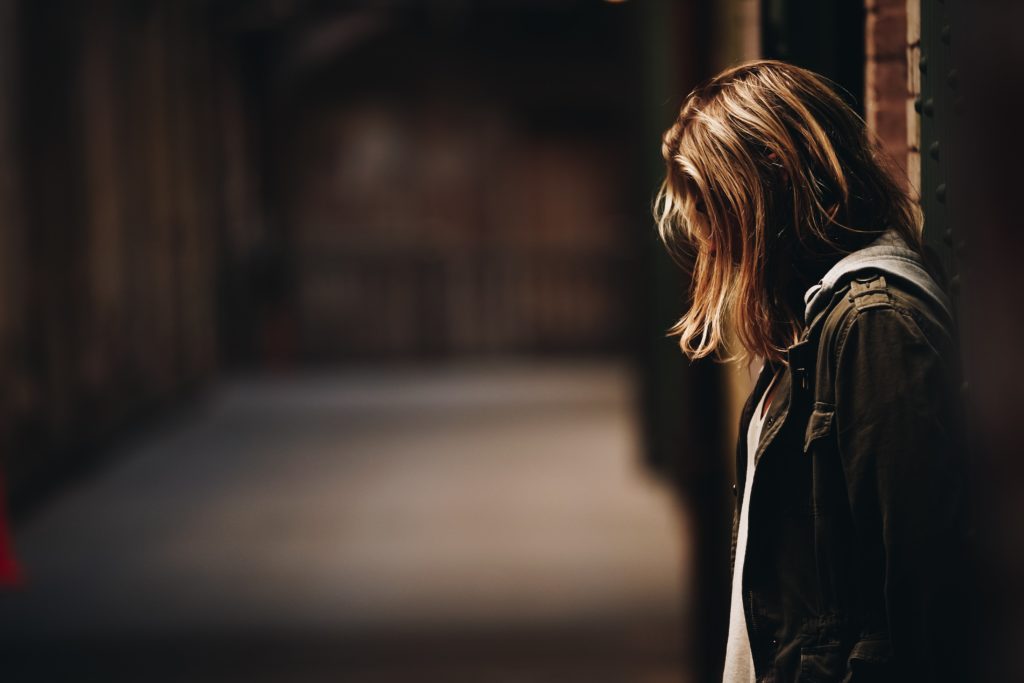 En jente står i profil med ryggen til en vegg langs en mørk gate. Hun har bøyd hodet og det halvlange blonde håret skjuler ansiktet hennes.