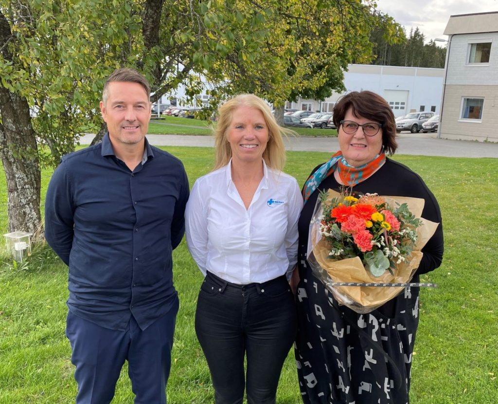 Tre personer står ved siden av hverandre og smiler. En mann og to kvinner. Hun ene holder en blomsterbukett i hånden. 