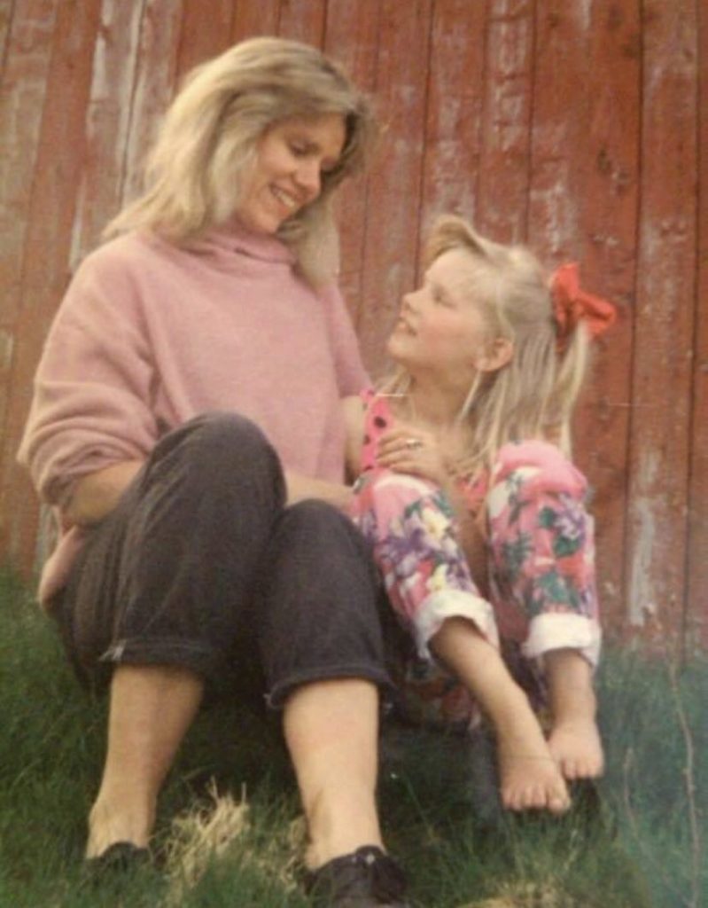 Camilla Dahle smiler opp mot sin mor der de sitter på gresset ved gården hun vokste opp
