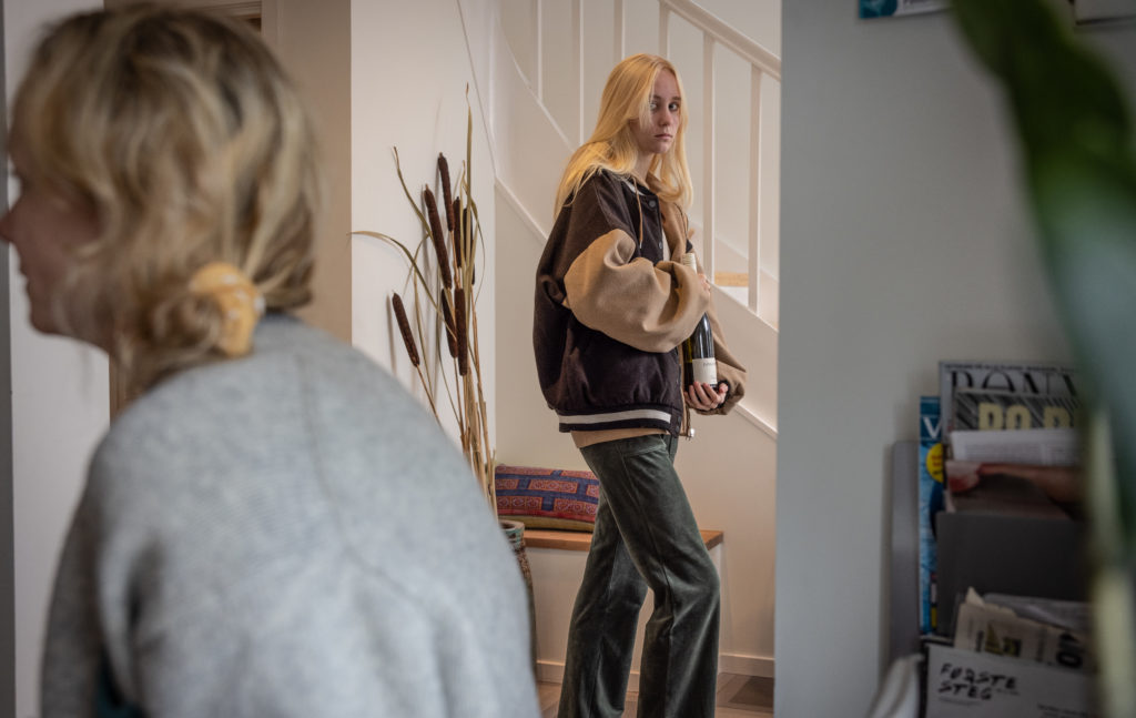 En tenåringsjente med blondt skulderlangt hår og et lurt blikk prøver å smugle en vinflaske ut av huset uten at moren ser det.