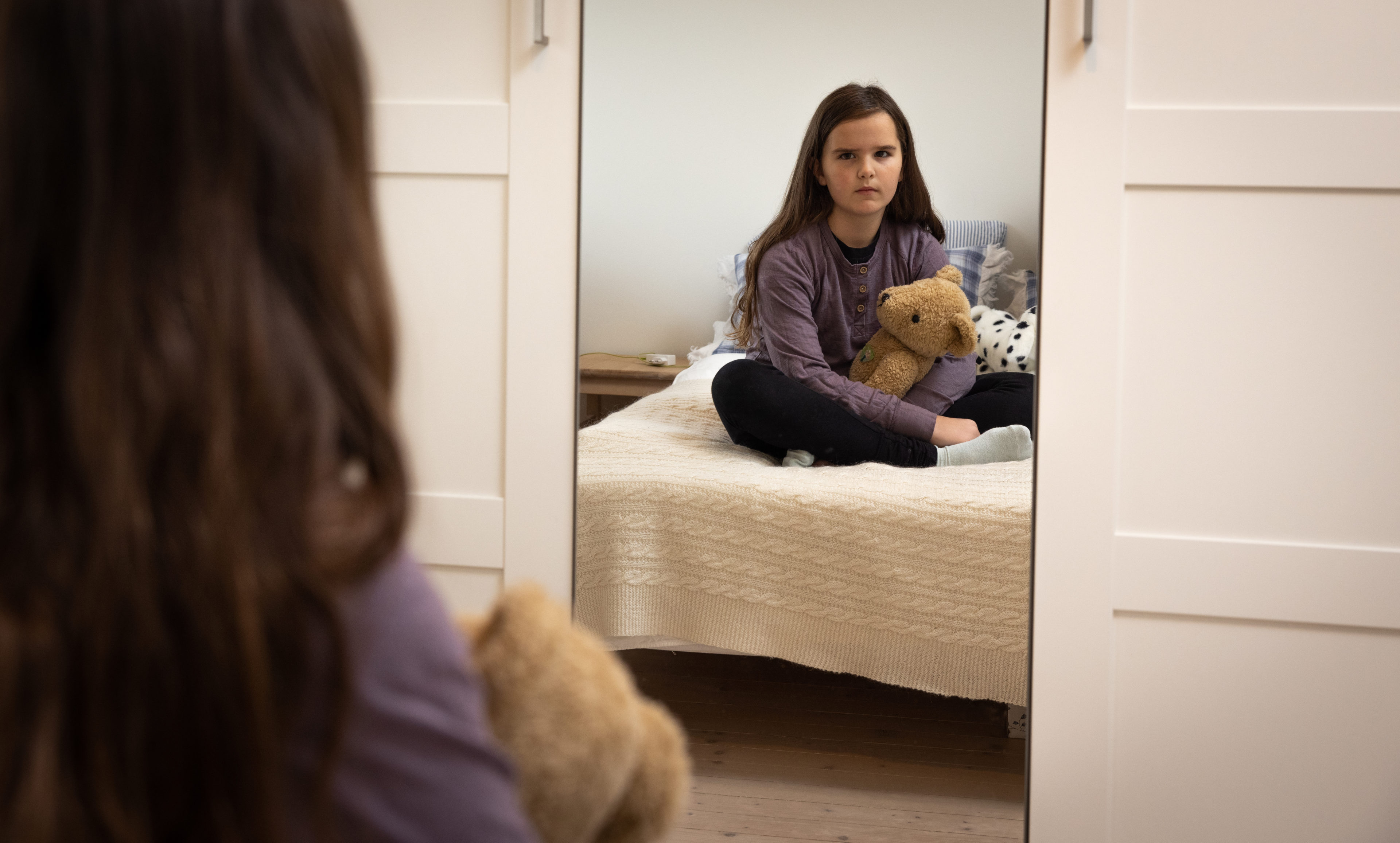En ung jente med lilla genser og brunt langt hår sitter på sengekanten med en beskyttende arm rundt bamsen mens hun stirrer alvorlig inn i speilet.
