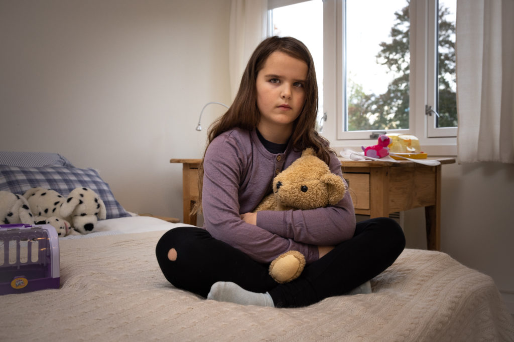 En ung jente med lilla genser og brunt langt hår sitter på sengekanten med en beskyttende arm rundt bamsen mens hun sitter alvorlig inn i kameraet.