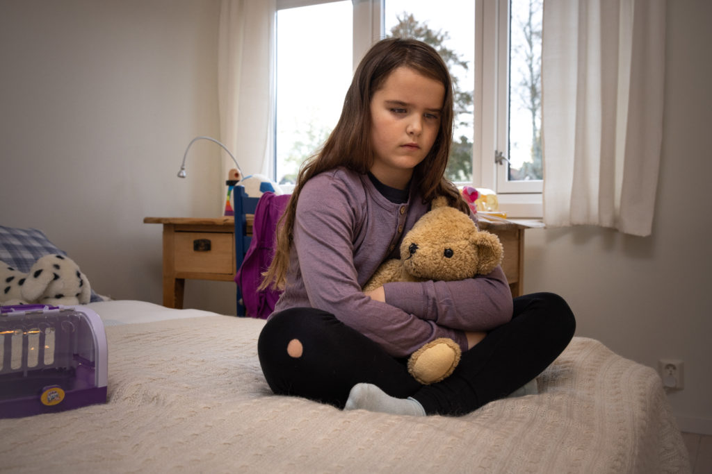 En ung jente med lilla genser og brunt langt hår sitter på sengekanten med en beskyttende arm rundt bamsen mens hun sitter alvorlig ned i gulvet.