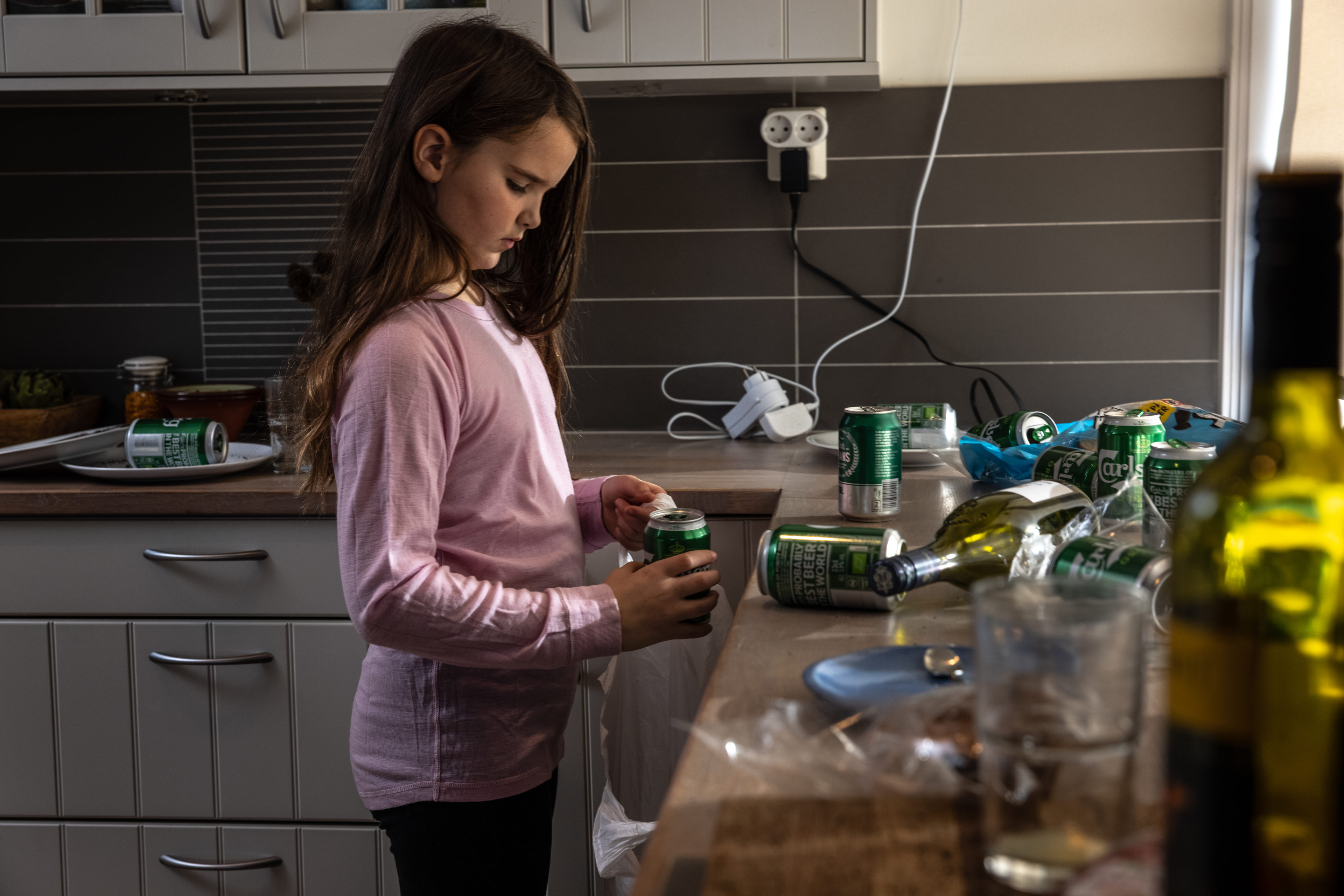 En liten jente står på kjøkkenet med et alvorlig blikk mens hun prøver å rydde ølboksene som flyter på kjøkkenbenken.
