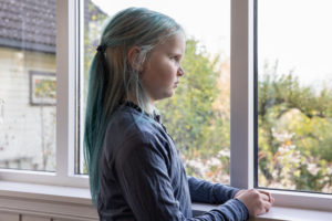 En jente i 10-årsalderen med blått hår stirrer alvorlig ut vinduet hjemme.
