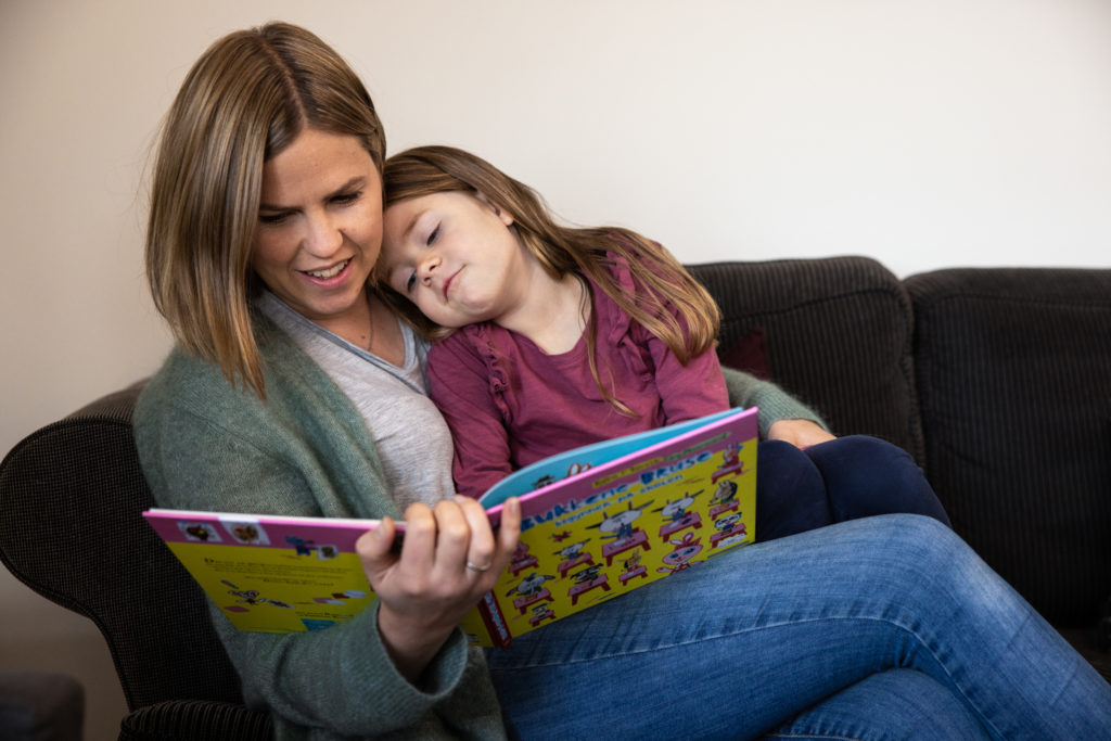 En mor sitter i sofaen med sin lille datter på fanget mens hun leser en barnebok.