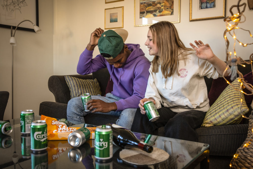 Et ungt par sitter i sofaen og drikker øl. Det er flere ølbokser på bordet. Paret smiler og det er god feststemning.