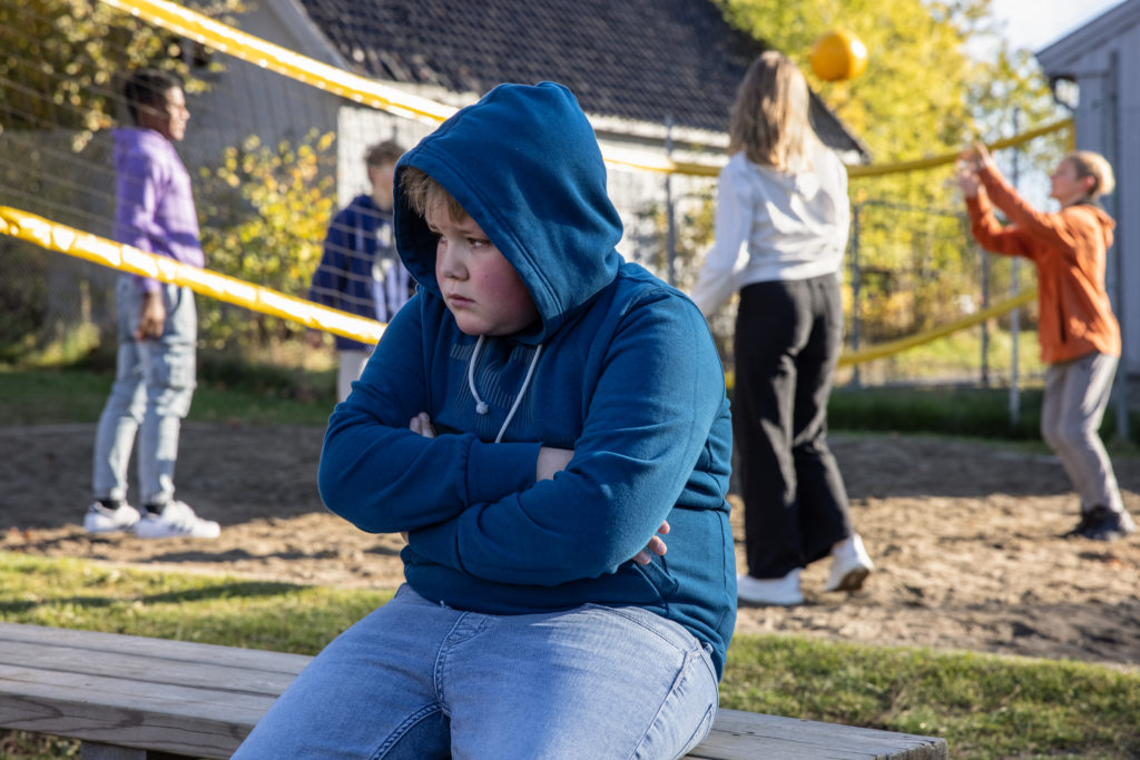 En gutt sitter på en benk med armene i kors. Han ser trist ut