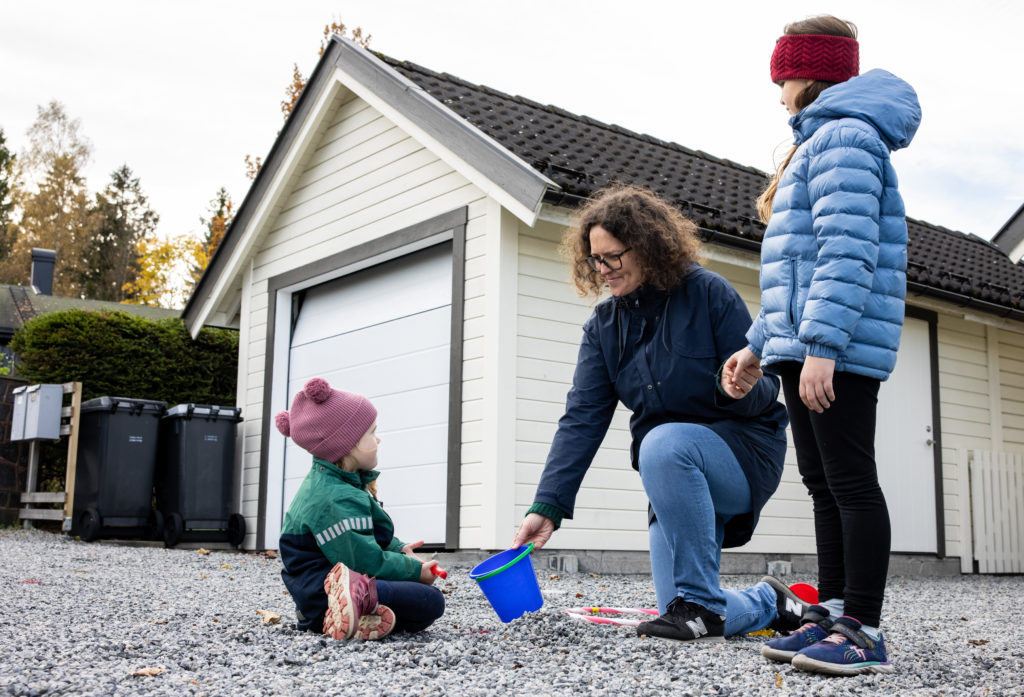 En liten jente sitter på bakken. En voksen dame sammen med et barn tilbyr den lille jenta en bøtte. Den voksne smiler