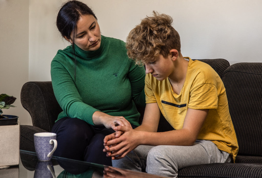 En mor trøster sønnen som sitter ved siden av henne i sofaen og er lei seg