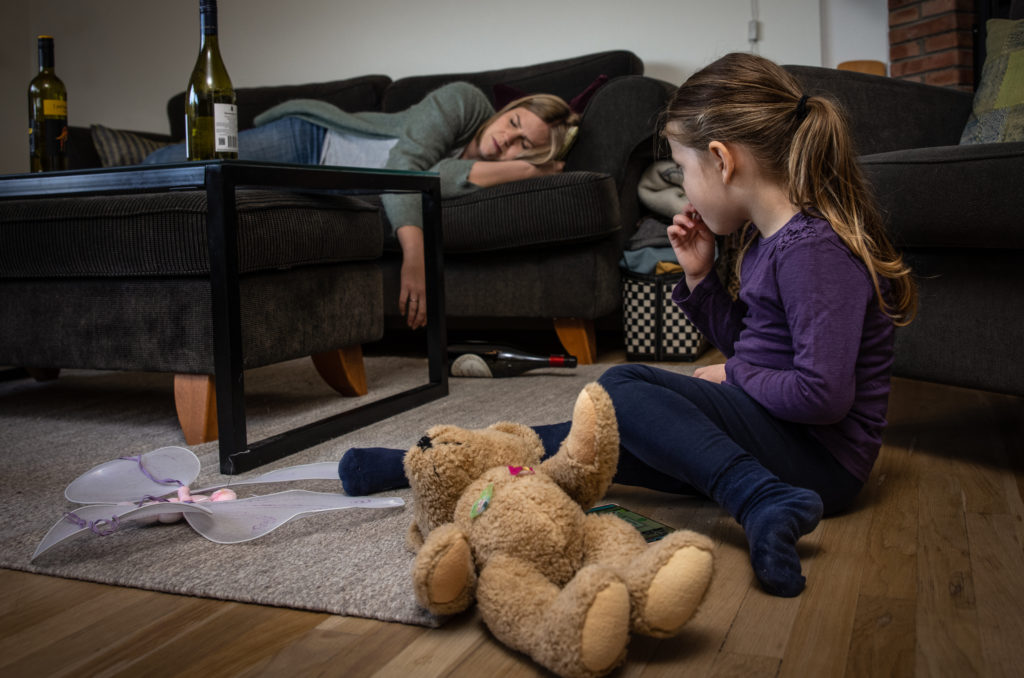 En jente sitter på gulvet og ved siden av bamsen sin og ser bort på moren sin. Hun er lei seg og alene. Moren hennes ligger og sover på sofaen og man ser vinflasker på bordet