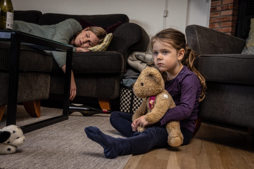 En jente sitter på gulvet og holder bamsen sin i armene. Hun er lei seg. Moren hennes ligger og sover på sofaen og man ser vinflaske på bordet