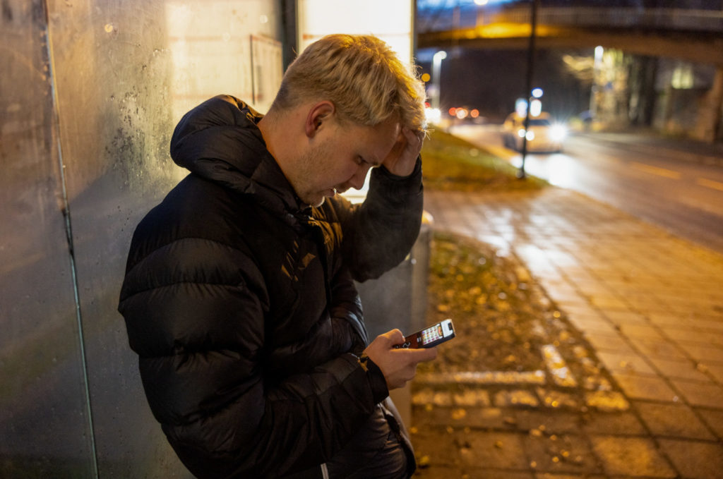 En mann spiller på mobilen med et bekymringsfullt utrykk i ansiktet, mens han står og venter på bussen.