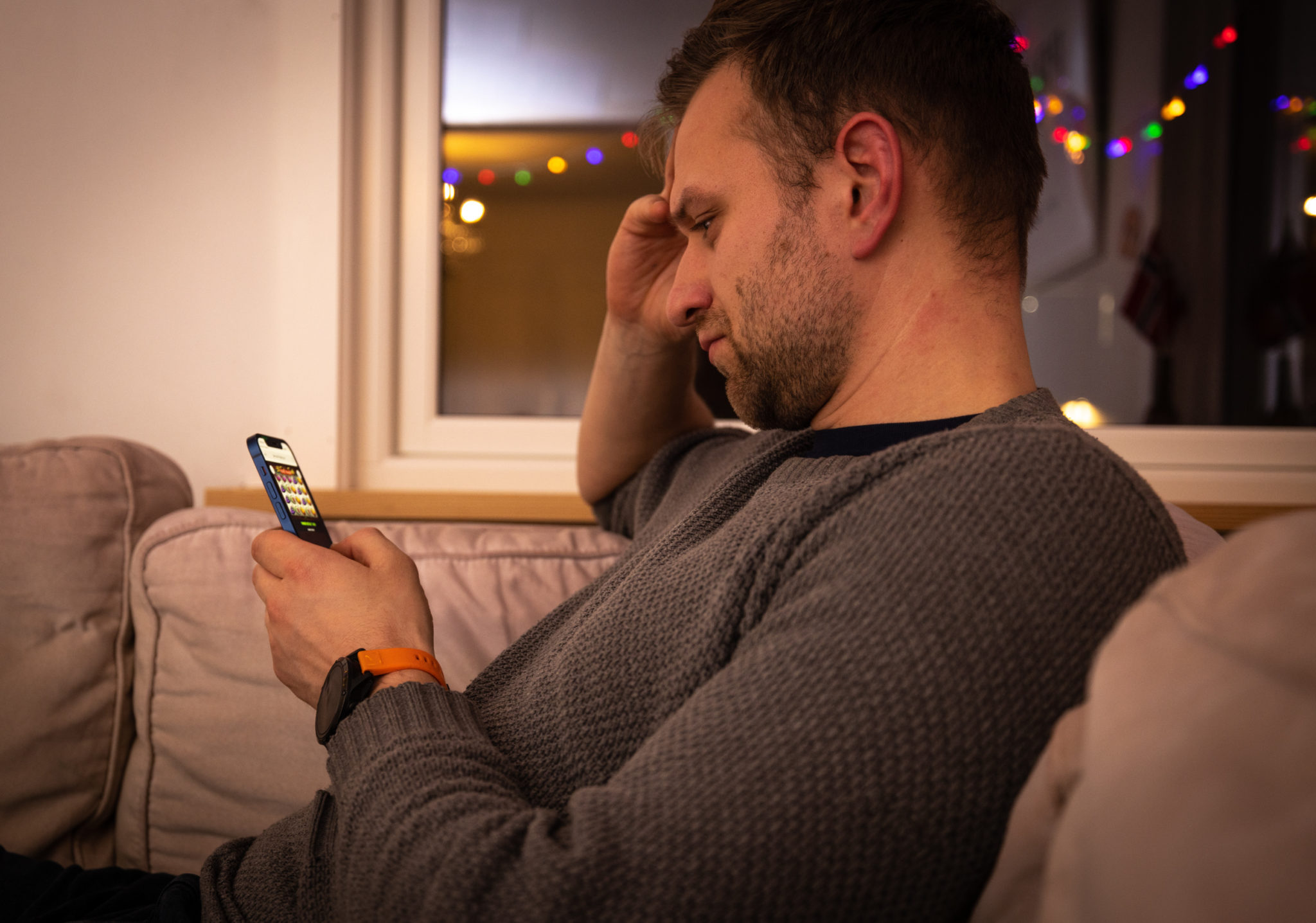 En mann sitter i sofaen og spiller pengespill på telefonen med et konsentrert utrykk i ansiktet.