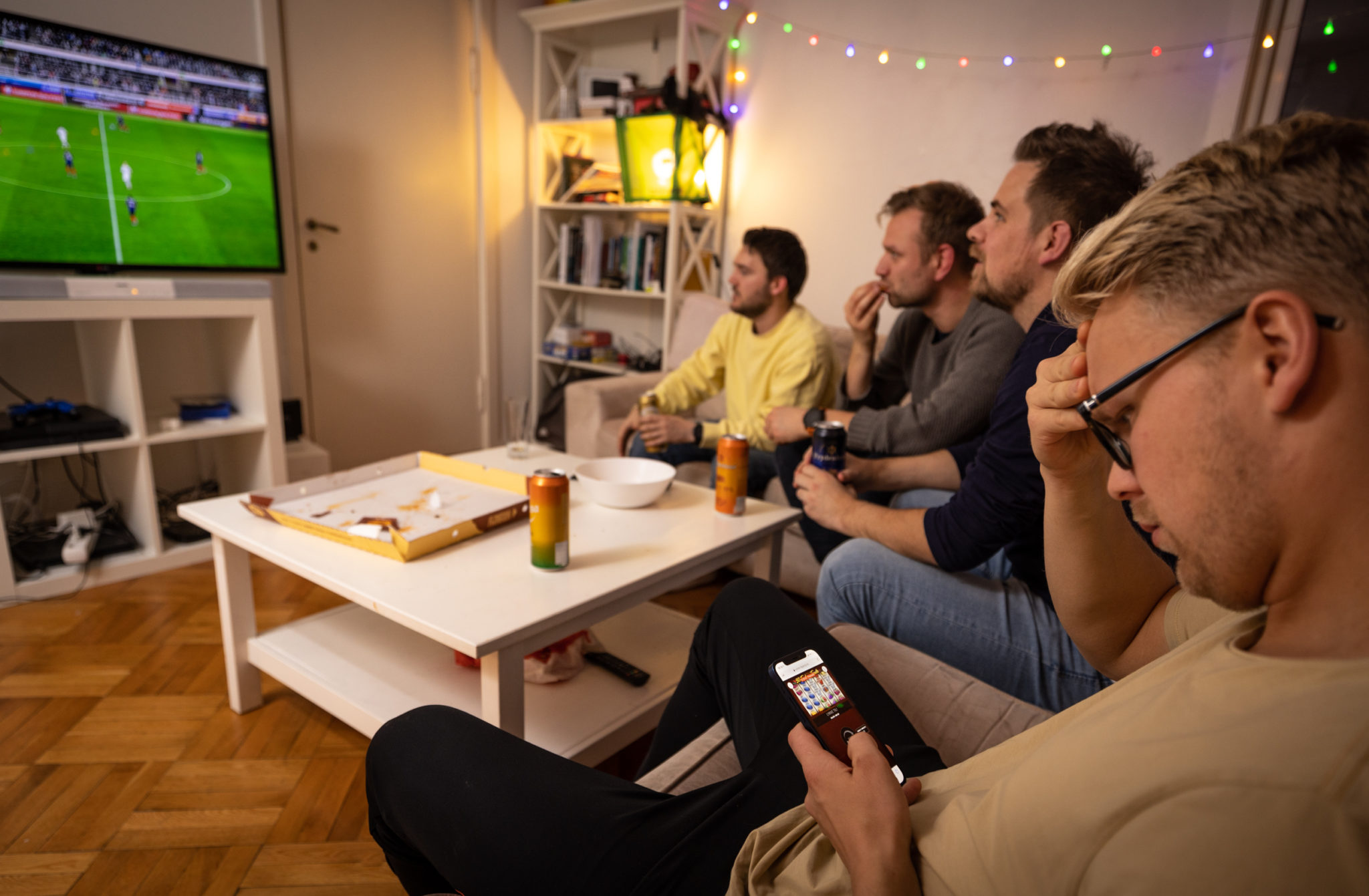 En guttegjeng sitter og ser på fotball på TV. En av dem sitter derimot på enden av sofaen og spiller pengespill på telefonen med et konsentrert utrykk i ansiktet.