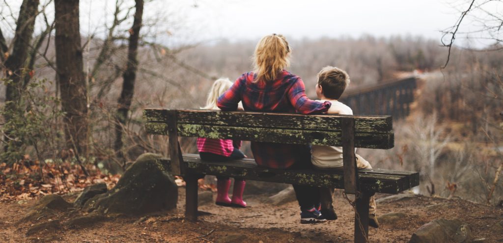 kvinne og to barn sitter med ryggen til på en benk i skogen