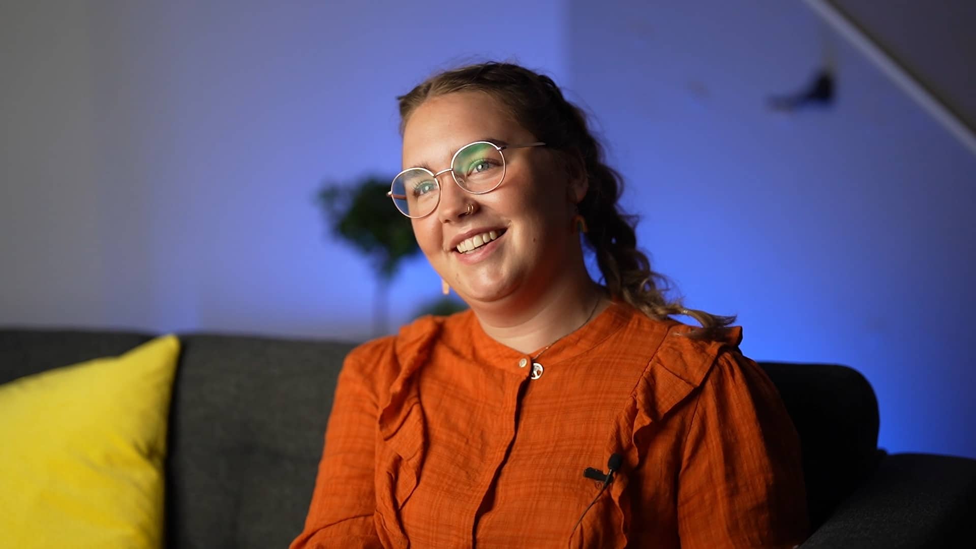 Sara er tidligere elev hos Blå Kors videregående skole Øvebrø. Kvinne med brune fletter, briller og oransje bluse smiler.