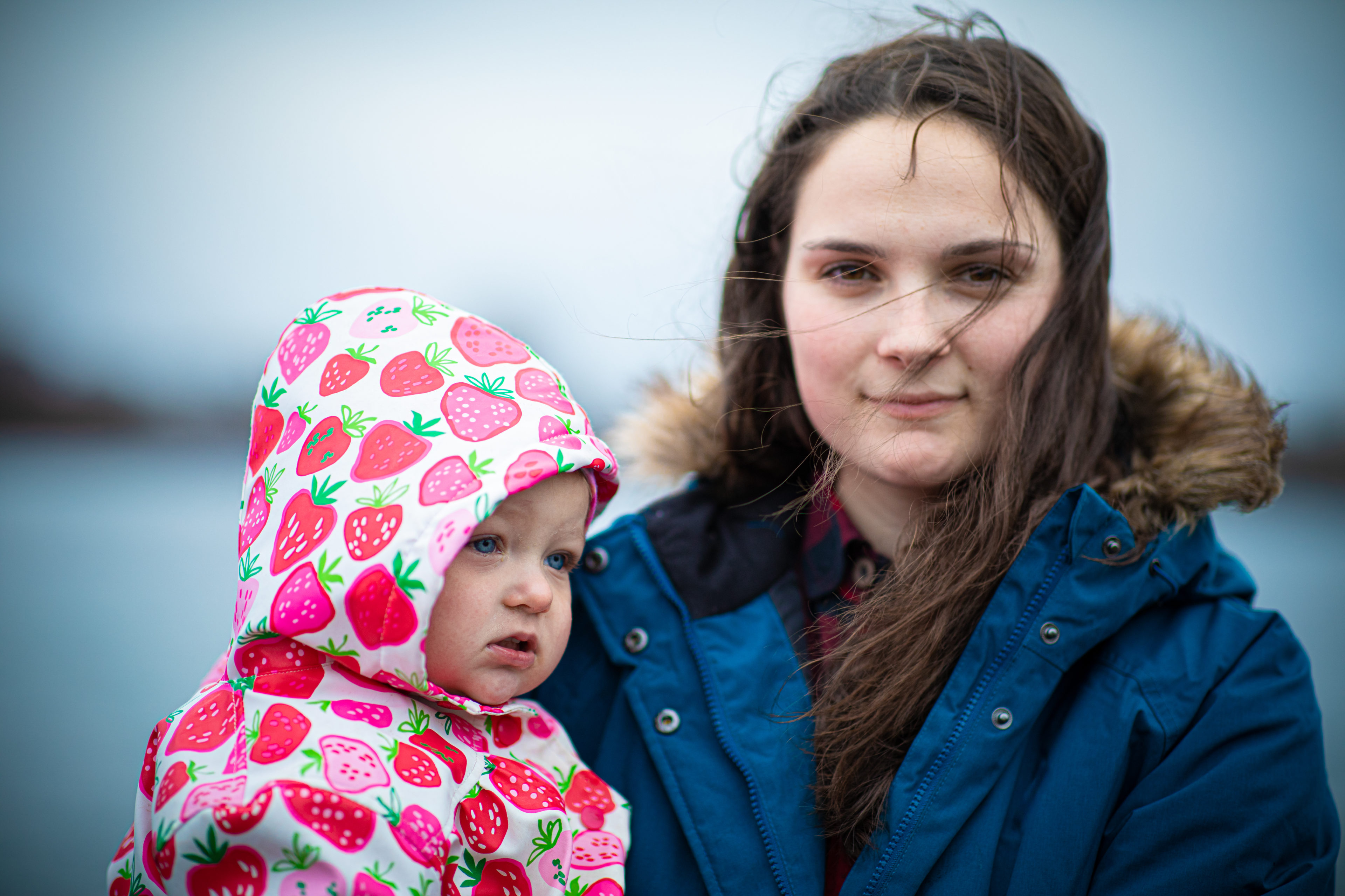 Bilde av en ung kvinne med brunt hår og blå jakke. Hun holder datteren sin i armene. Datteren er rundt 4 år, og har på seg en rosa regnjakke med røde prikker.