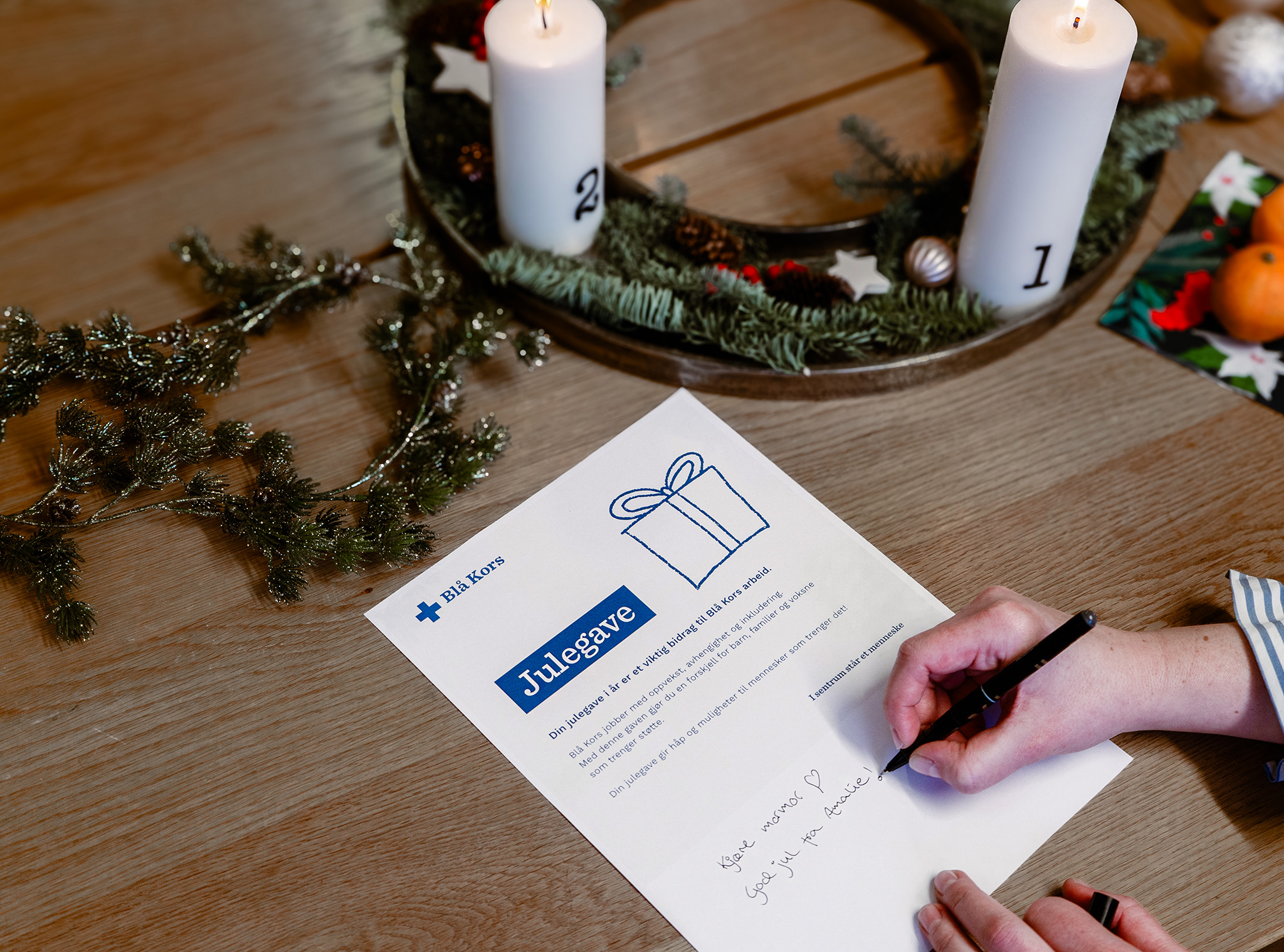 Bord er dekket med juledekorasjoner og en kvinne skriver på julegave med mening gavekortet som hun gir fra Blå Kors til sin mormor. "God jul mormor. Hilsen Amalie".