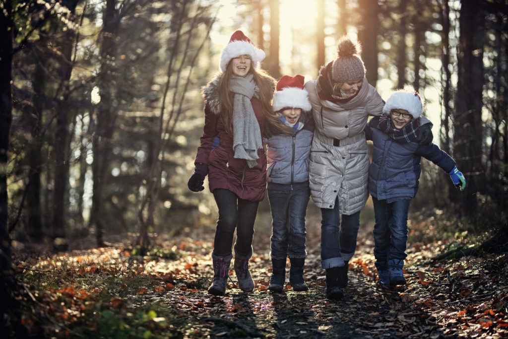 En mor på tur i skogen med sine tre barn. De smiler og koser seg sammen. Barna har på seg nisseluer og vinterklær.