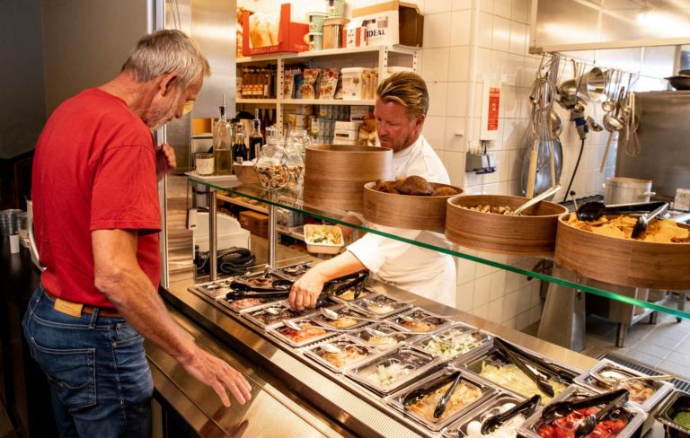 En mann står ved buffe og skal velge seg noe å spise. Det er fra en salatbar. Det er også en kokk som står der og viser han