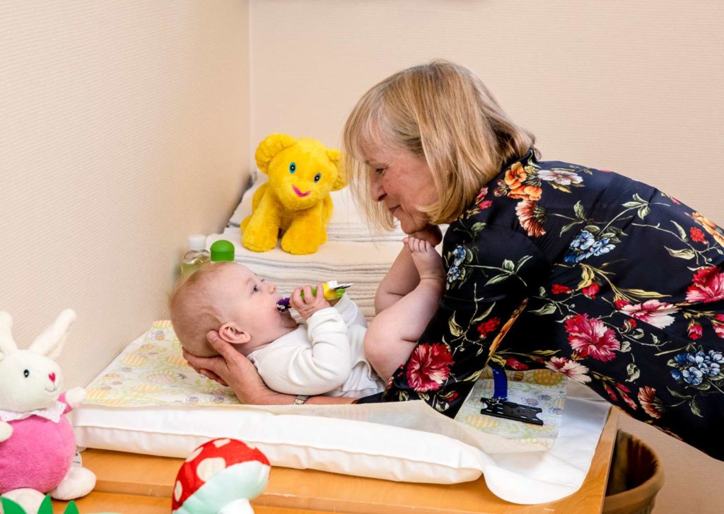 En baby ligger på en stellemappe og blir løftet litt opp av en kvinne. Hun smiler og duller med barnet