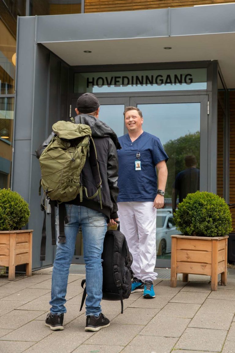 Utenfor hovedinngangen Blå Kors Lade Behandlingssenter er det en behandler fra Klinikken som tar imot en pasient. Pasienten kommer i en grønn ryggsekk og svarte joggesko